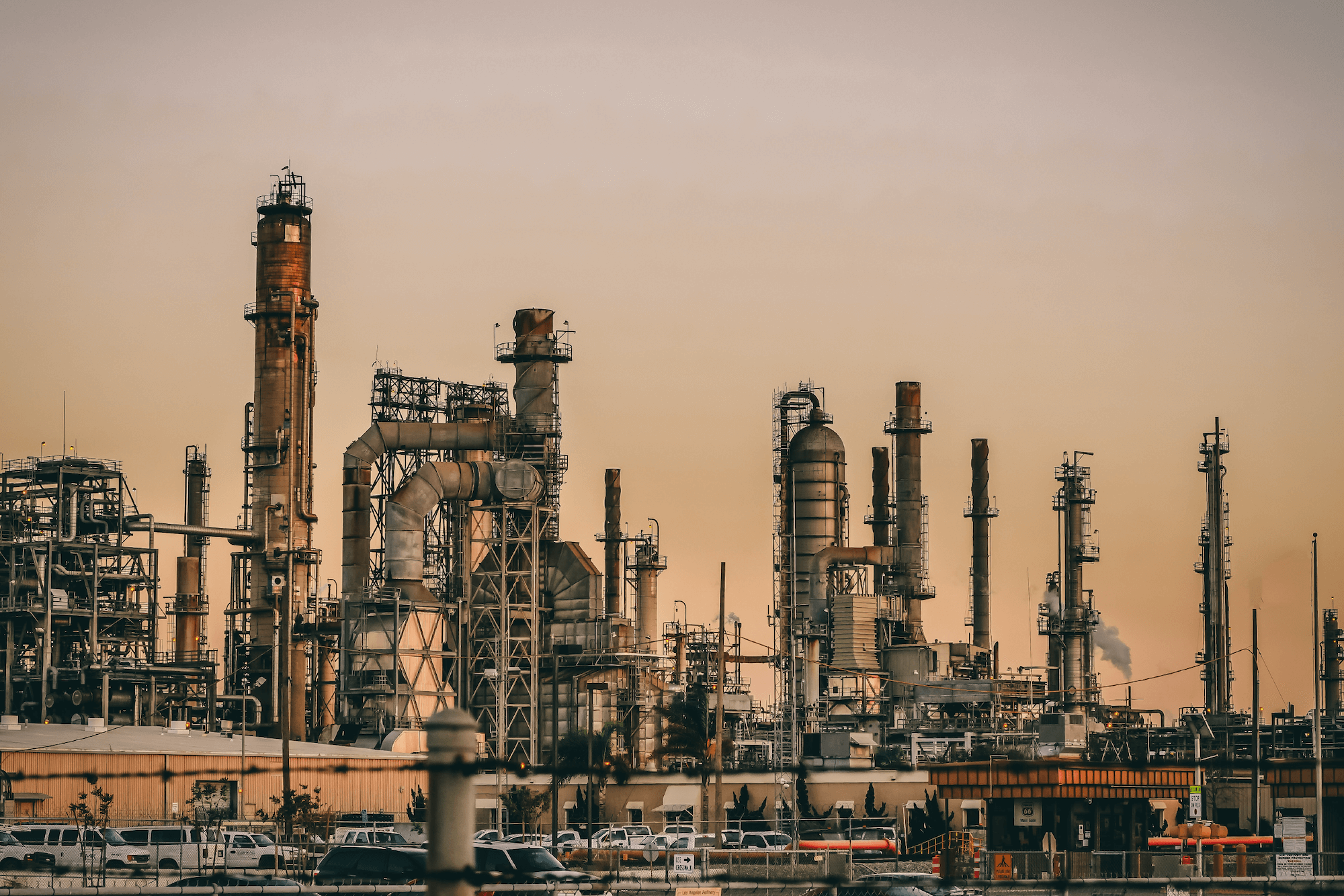 An industrial complex at sunset with intricate piping and multiple smokestacks releasing steam against a dusky sky.